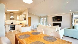 A modern living room with a wooden dining table, a sofa, and a TV mounted on the wall. - Host & Stay | GRE06 Deneb Lodge (Amotherby)