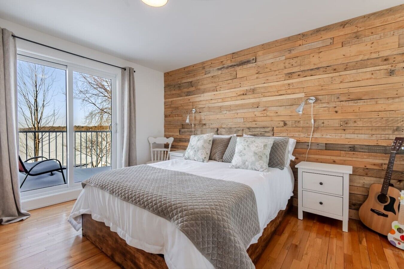 Une chambre avec un mur en bois, un lit avec des oreillers, une table de chevet, une guitare et un balcon avec des chaises.