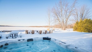 Un bain à remous entouré de chaises et de tables recouvertes de neige, avec vue sur un lac et des arbres en arrière-plan.