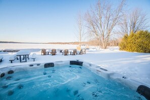 Un bain à remous entouré de chaises et de tables recouvertes de neige, avec vue sur un lac et des arbres en arrière-plan.