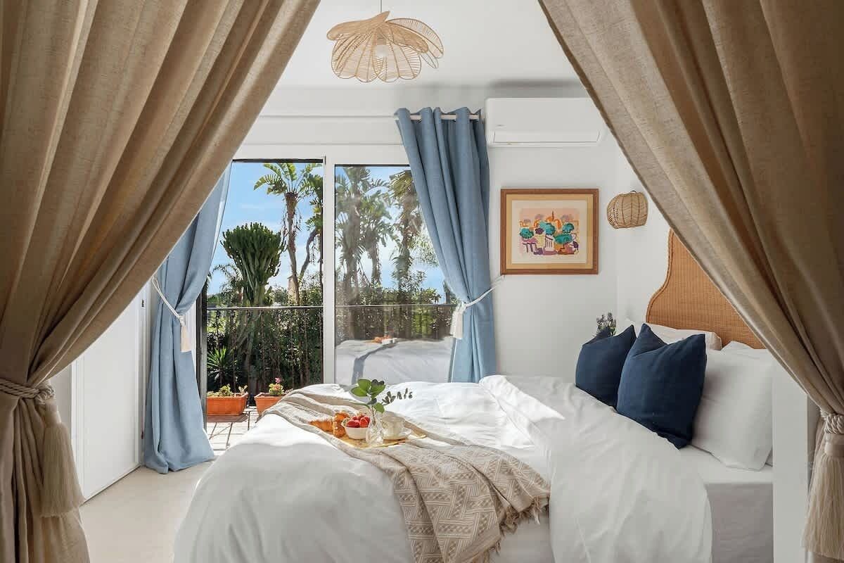 A bedroom with a bed, curtains, a painting, and a view of palm trees.