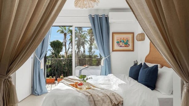 Une chambre à coucher avec un lit, des rideaux, un tableau et une vue sur des palmiers.