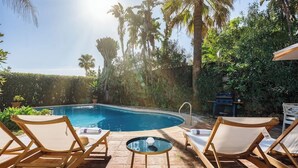 Un espace aménagé au bord de la piscine, avec des chaises longues, une table en verre et une piscine entourée d’une végétation tropicale.