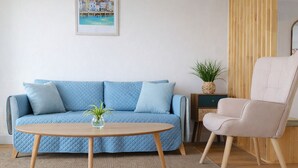 Ein Wohnzimmer mit einem blauen Sofa, einem runden Holztisch, einem beigen Sessel, einem kleinen Beistelltisch mit Pflanze und einem gerahmten Bild an der Wand.