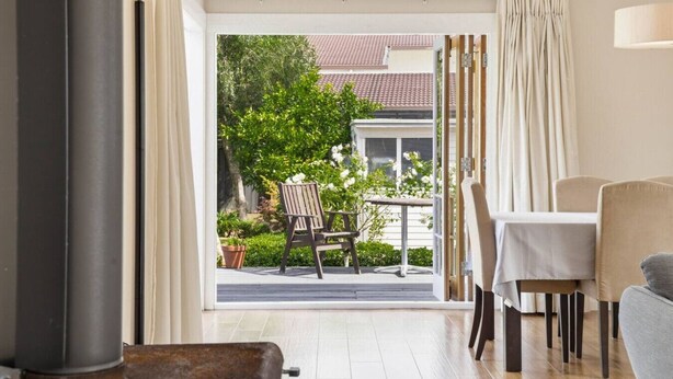 A room with a dining table, chairs, a sofa, and a view of a garden and neighboring buildings. - 1920s Character Gem in Sandringham | Garden & Deck (Auckland Central)