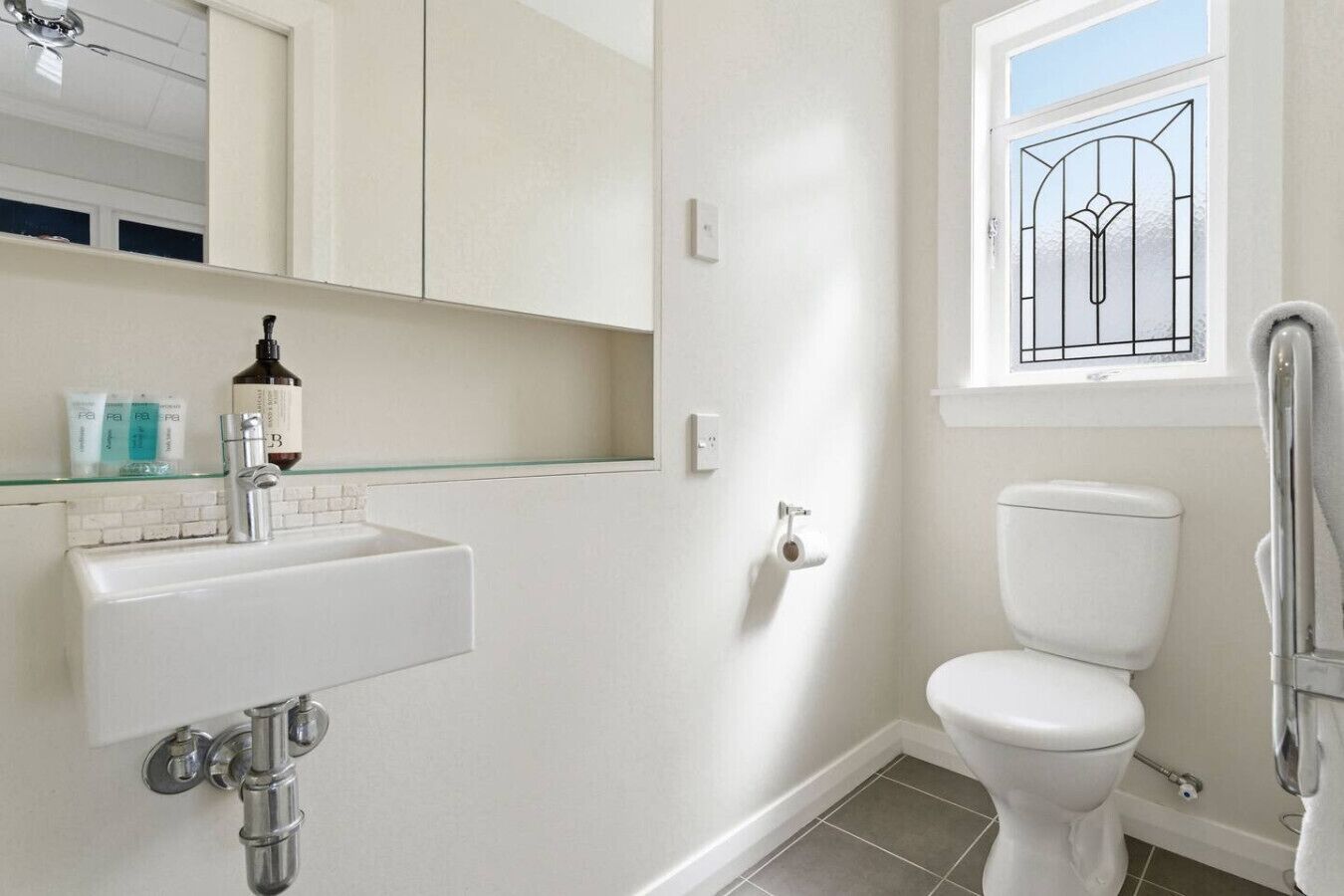 Une salle de bain avec un lavabo blanc, un miroir, des toilettes et une fenêtre ornée d’un motif décoratif.