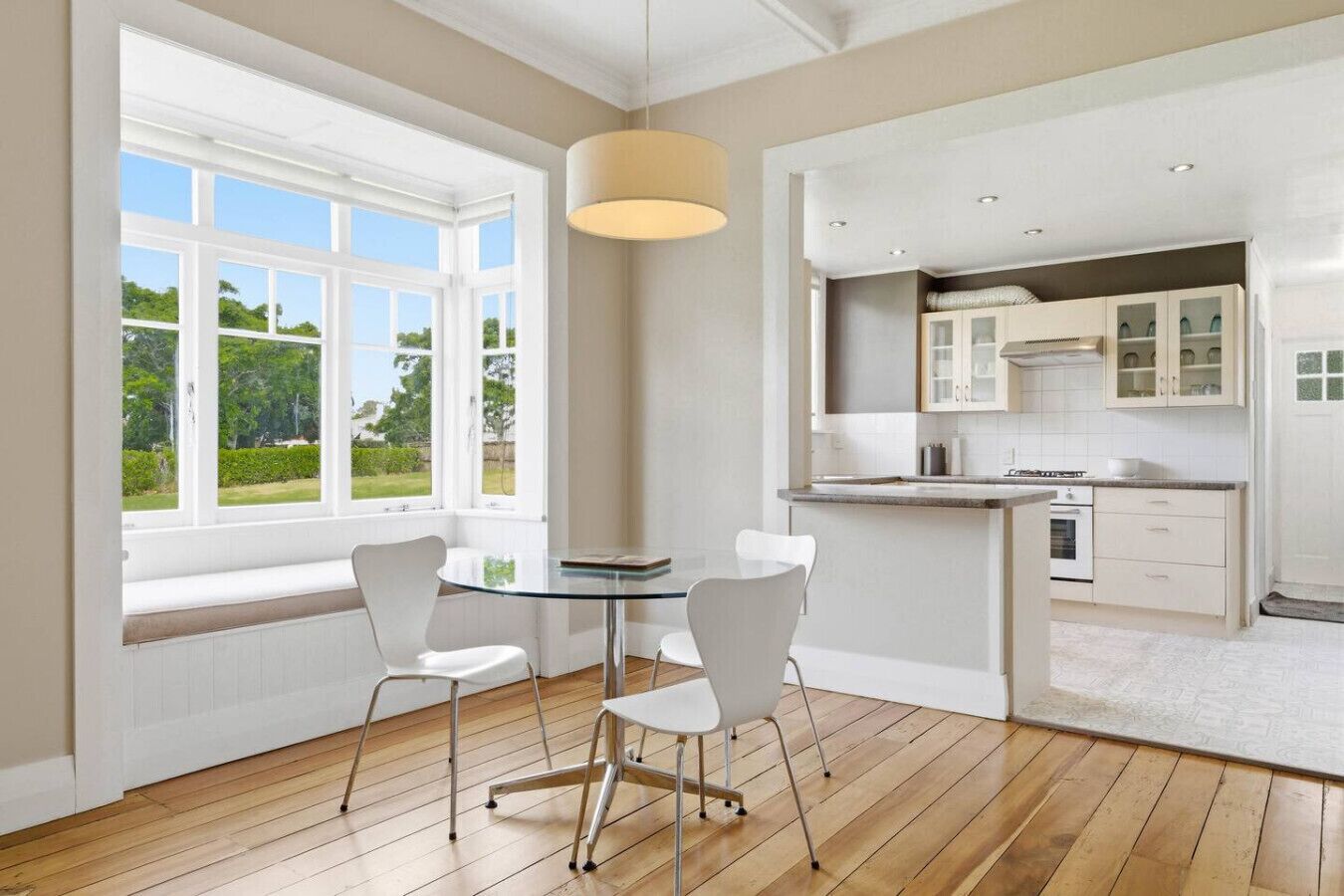 A modern kitchen with white cabinetry, a dining area with a glass table and chairs, and large windows with a view of greenery.