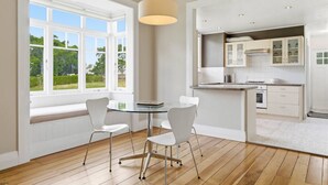 A modern kitchen with white cabinetry, a dining area with a glass table and chairs, and large windows with a view of greenery. - 1920s Character Gem in Sandringham | Garden & Deck (Auckland Central)