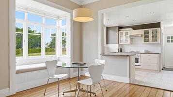 A modern kitchen with white cabinetry, a dining area with a glass table and chairs, and large windows with a view of greenery.
