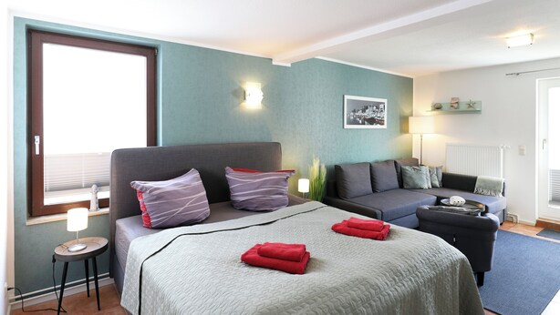A modern hotel room with a bed, a sofa, a small table, and a window with blinds. - Appartement Nr. 05 (Rostock)