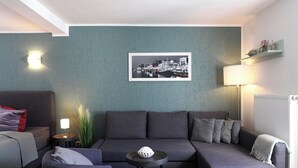 A modern living room with a grey sofa, a small round table with a lamp, a framed cityscape picture on the wall, and a radiator. - Appartement Nr. 05 (Rostock)