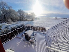 Een besneeuwde buitenruimte met een bank en tafels, een hekwerk en uitzicht op een waterpartij.