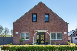 Een rode bakstenen woning met een puntig dak, twee ramen, een zwarte deur en een klein portaal met potplanten.
