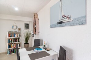 Un comedor con una mesa blanca, sillas negras, una estantería con libros, una planta en maceta y un cuadro de un velero en la pared.