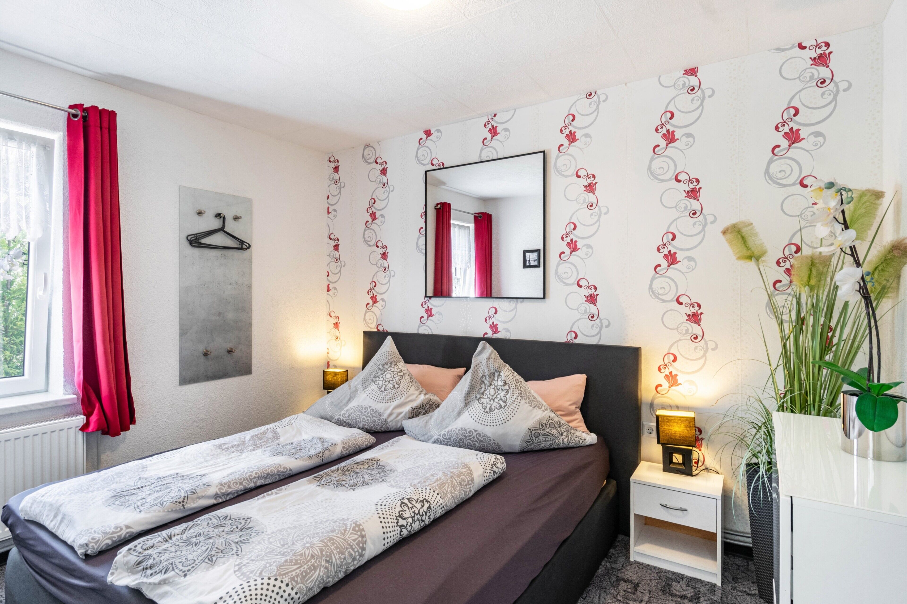 Ein Schlafzimmer mit Bett, Nachttischen, einem Spiegel, einem Fenster mit Vorhängen und einer Wand mit einem Blumenmuster.
