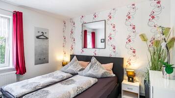 Ein Schlafzimmer mit Bett, Nachttischen, einem Spiegel, einem Fenster mit Vorhängen und einer Wand mit einem Blumenmuster.