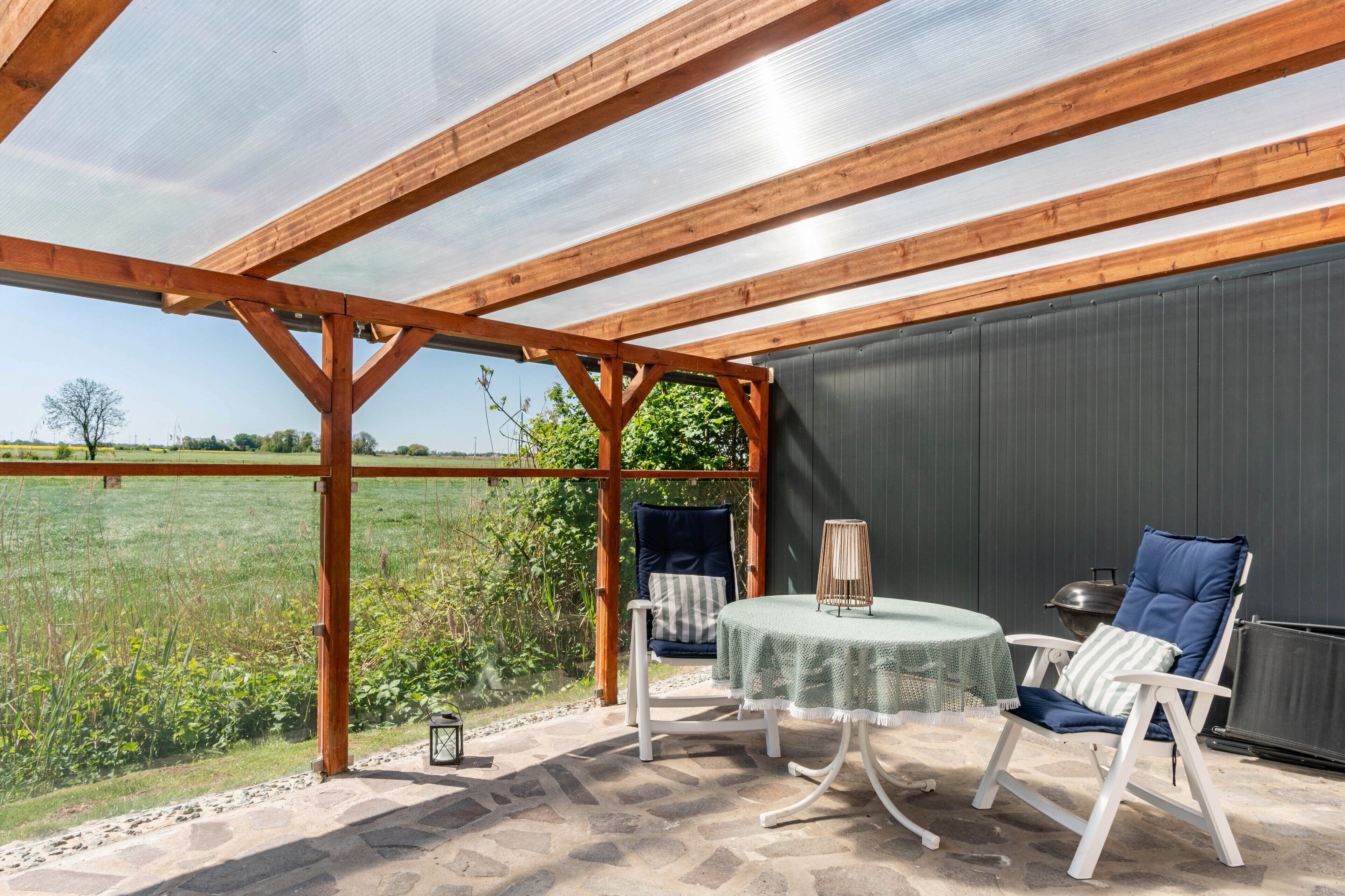 Eine überdachte Terrasse mit Tisch und Stühlen, Blick auf ein Feld und ein Metallgrill.