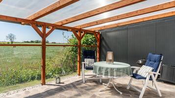 Eine überdachte Terrasse mit Tisch und Stühlen, Blick auf ein Feld und ein Metallgrill.