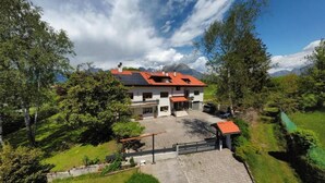 Una casa de dos pisos con techo rojo, rodeada de follaje y una montaña al fondo.