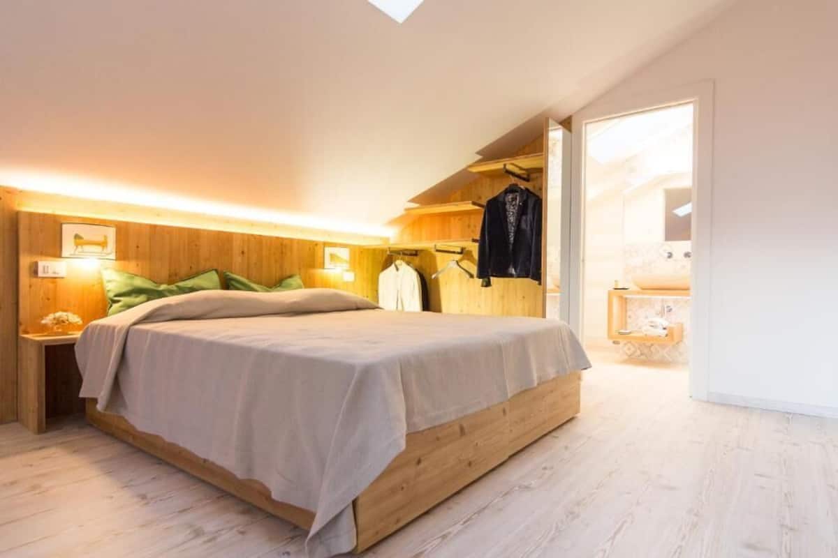 A bedroom with a wooden bed frame, a nightstand with a lamp, and a wardrobe with clothes hanging inside.