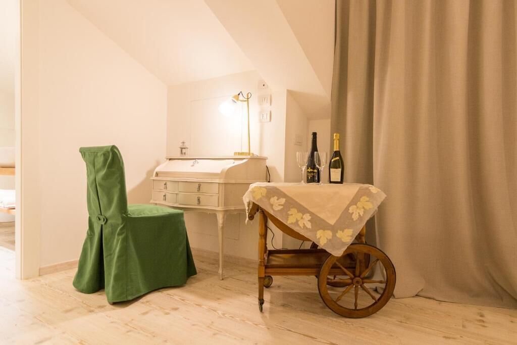 Une pièce avec un plancher en bois, une chaise verte, une petite table recouverte d’une nappe à motifs floraux, un chariot en bois, une commode blanche et une bouteille de vin posée sur la table.
