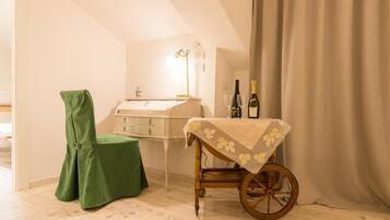Une pièce avec un plancher en bois, une chaise verte, une petite table recouverte d’une nappe à motifs floraux, un chariot en bois, une commode blanche et une bouteille de vin posée sur la table.