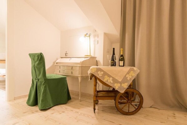 Una habitación con piso de madera, una silla verde, una mesita con mantel floreado, un carrito de madera, un aparador blanco y una botella de vino sobre la mesa.
