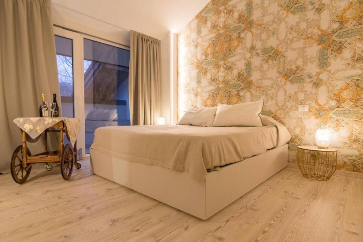 A bedroom with a large bed, a small table with bottles, a wheelchair, and a patterned wall.