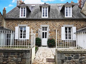 Una casa de piedra con puerta blanca, dos ventanas con contraventanas y un pequeño jardín con un arbusto de flores rojas.