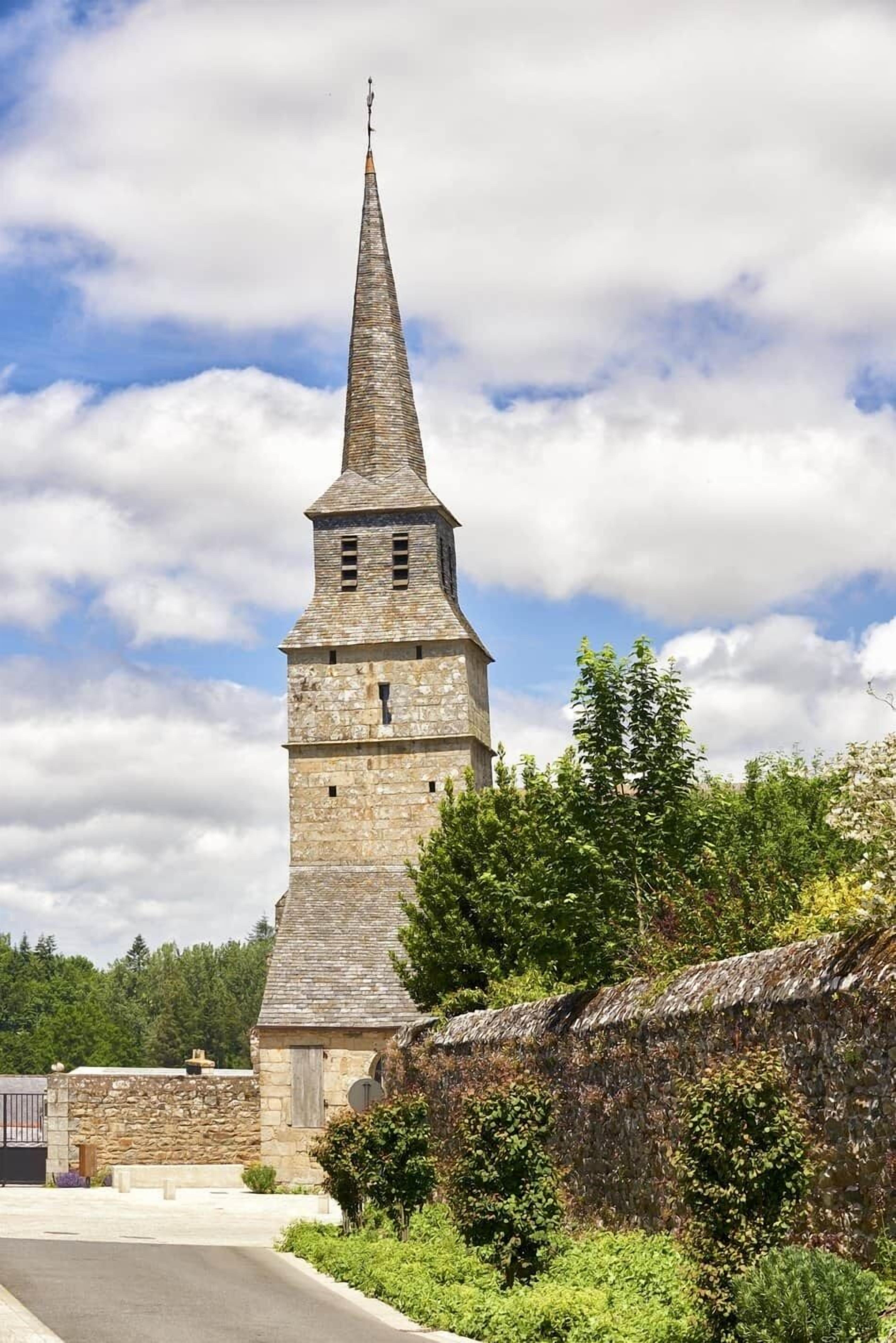 Una chiesa in pietra con un campanile slanciato, una strada pavimentata e un muro di pietra con vegetazione.