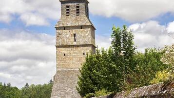 Una chiesa in pietra con un campanile slanciato, una strada pavimentata e un muro di pietra con vegetazione.
