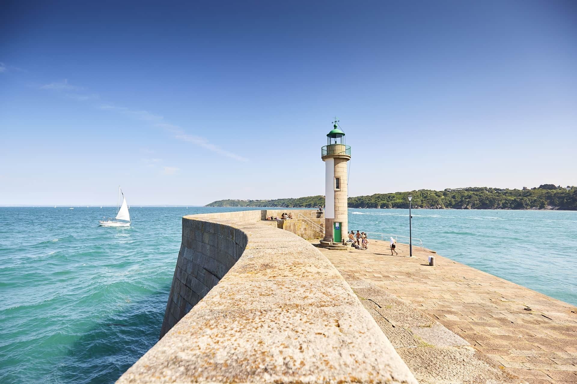 Un phare au bord de la mer, une promenade en béton et un voilier à l’horizon.