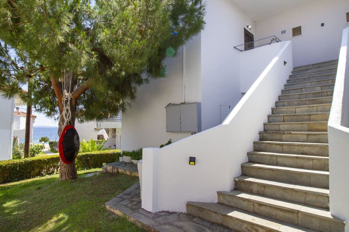 Ein weißes Gebäude mit einer Treppe, die zu einem Balkon führt, umgeben von Grün und Blick auf das Meer.