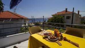 En balkon med et gult bord dækket til et måltid, med udsigt over et kystområde med bygninger og en klar himmel.