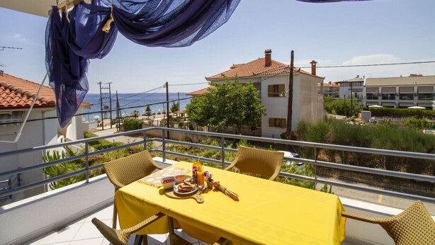 En balkon med et gult spisebord, der vender ud mod et kystlandskab med bygninger og en klar himmel.