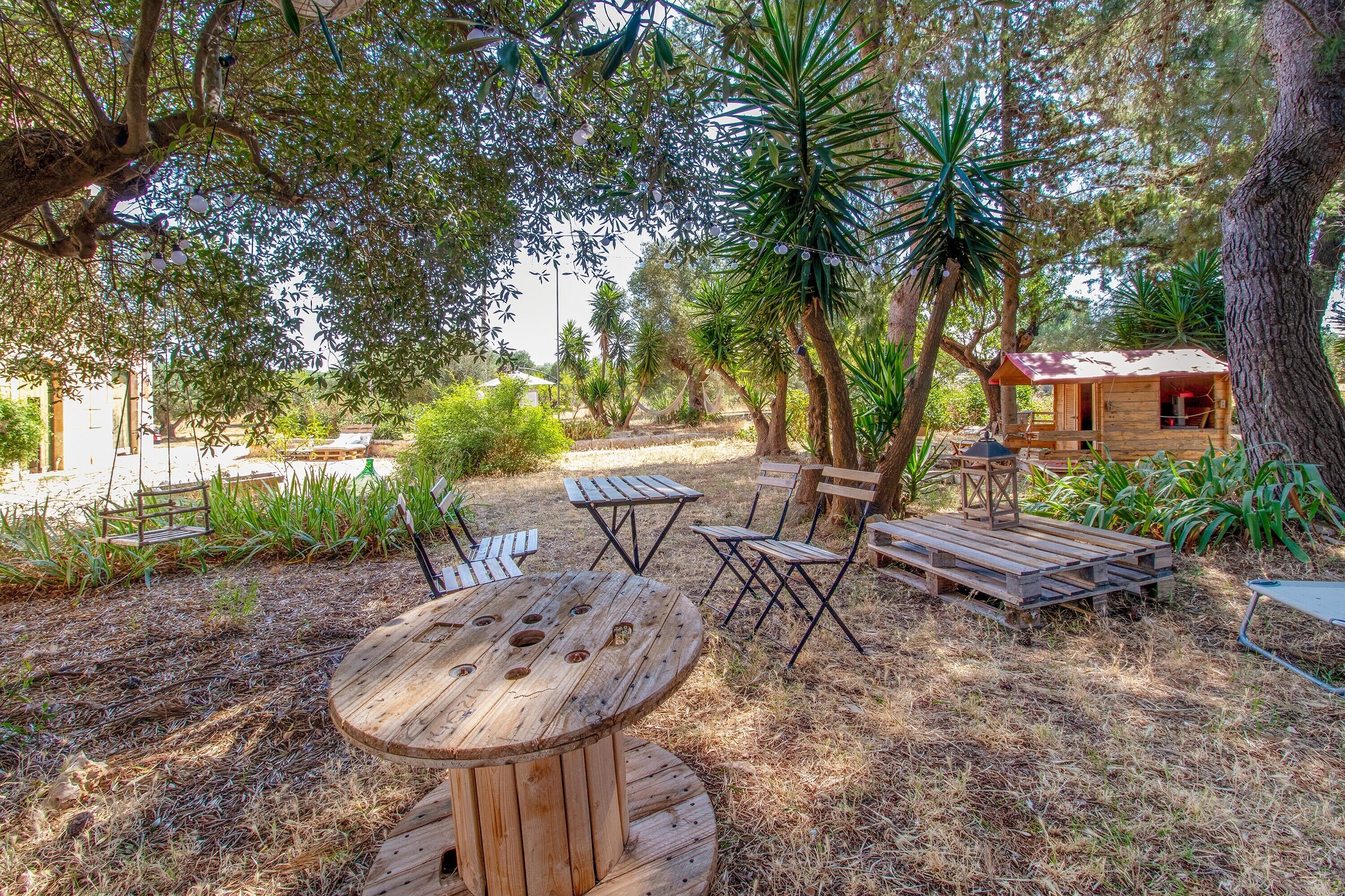 An outdoor seating area with wooden tables and chairs, surrounded by trees and a small wooden structure.
