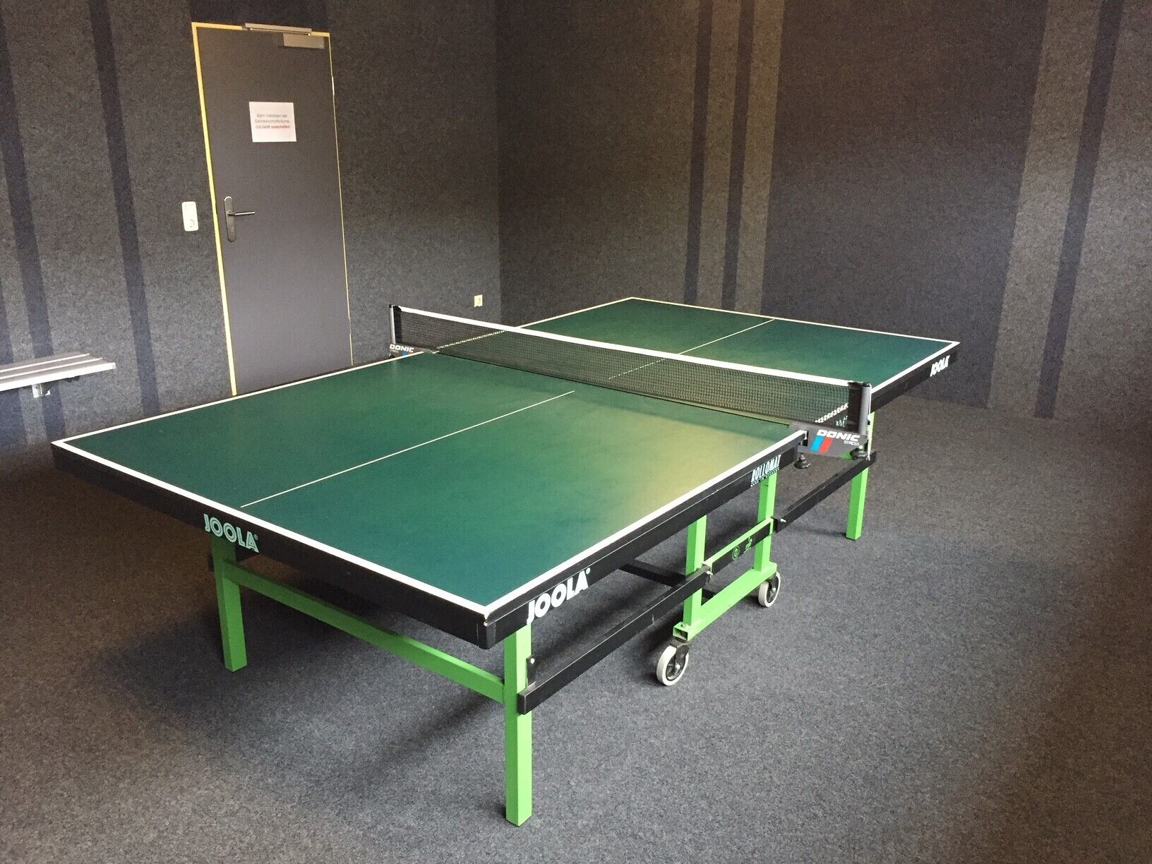 Une table de ping-pong aux plateaux verts et noirs, placée dans une pièce dont le sol est recouvert d’une moquette grise et les murs sont de couleur sombre.