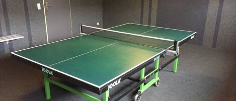 Une table de ping-pong aux plateaux verts et noirs, placée dans une pièce dont le sol est recouvert d’une moquette grise et les murs sont de couleur sombre.