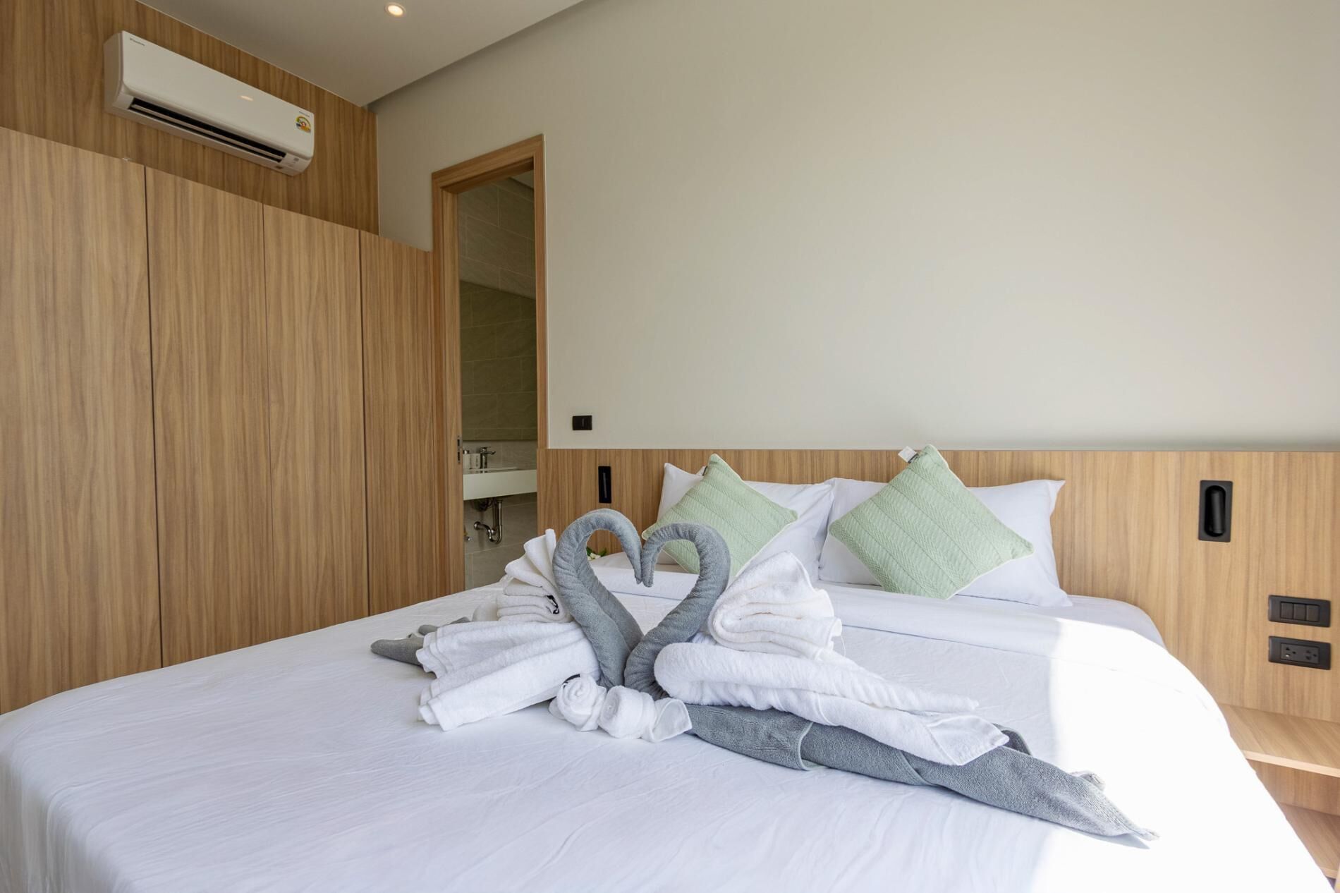 Quarto de hotel com cama, guarda-roupa de madeira, unidade de ar condicionado e banheiro visível por uma porta aberta.