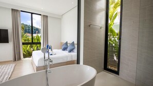 Ein modernes Hotelzimmer mit einer großen Badewanne, einem Bett mit blauen Kissen und Blick auf Grün durch große Fenster.