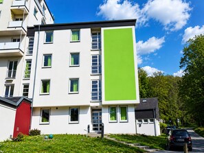 A multi-story building with a large green vertical panel, windows with green curtains, and a red door.