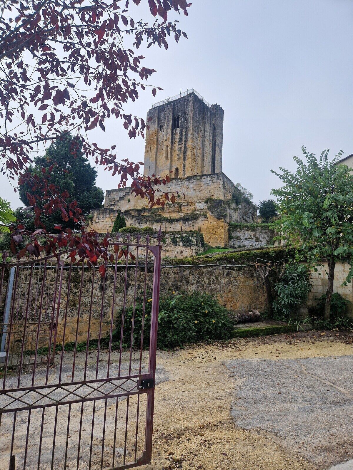 A historic stone building with a prominent tower, surrounded by a gravel area and a metal gate.