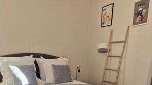 Una cama bien hecha con edredón gris y blanco, dos toallas blancas enrolladas y una escalera de madera apoyada en la pared.