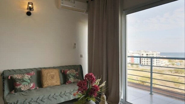 A living room with a sofa, a vase with flowers, and a view of buildings through a large window. - SeafrontView · Cozy T1 Apartment - 3 (Praia)