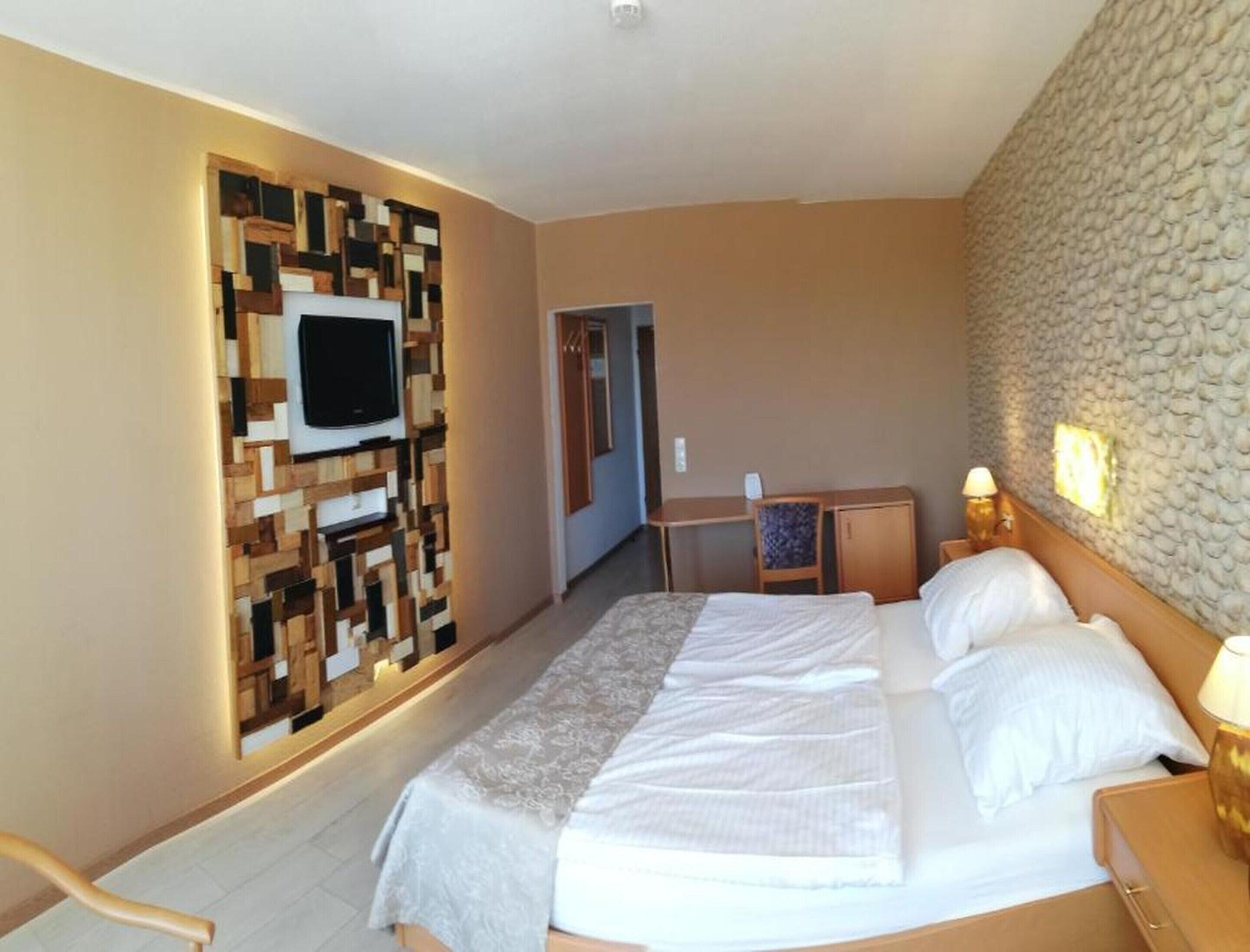 A hotel room with a bed, a desk, a chair, a television mounted on a wall, and a stone-textured wall.