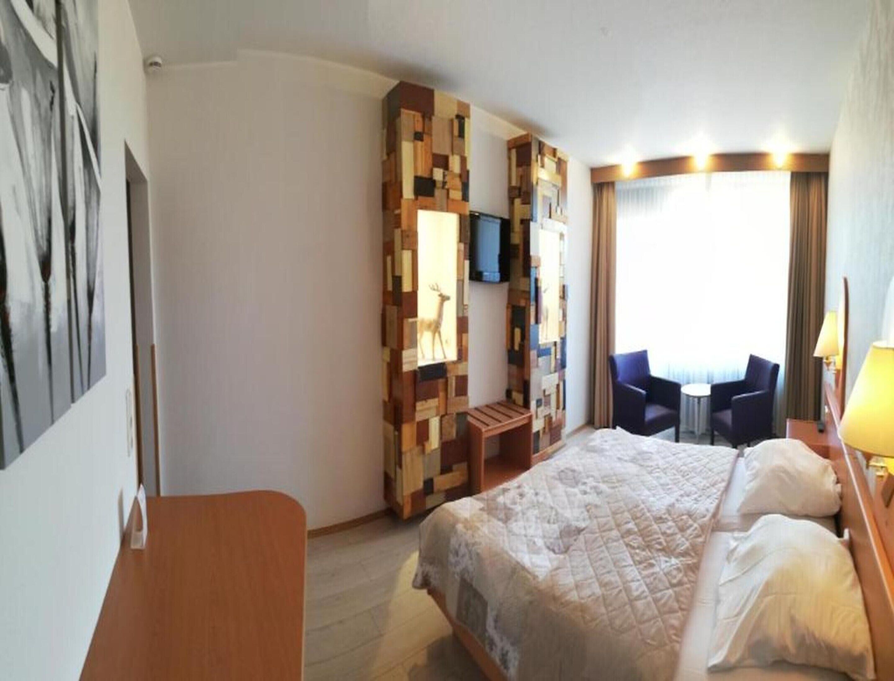 Quarto de hotel com cama, escrivaninha, cadeira, televisão e janela com cortinas.
