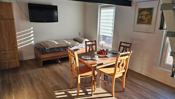 A compact room with a bed, a dining table with chairs, a TV mounted on the wall, and a window with blinds.