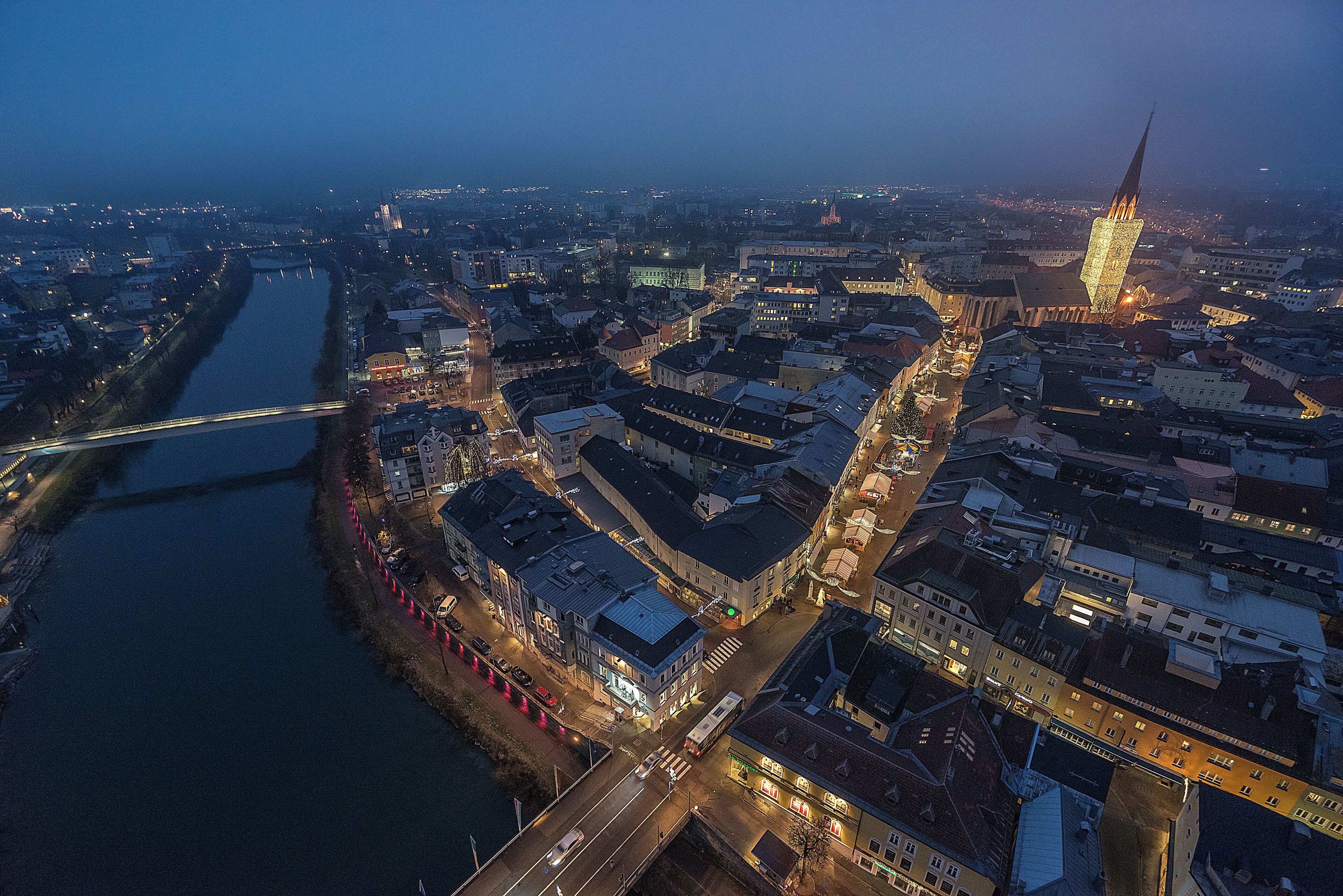 Luchtbeeld van een stadsgezicht in de schemering met een rivier, bruggen en verlichte gebouwen.