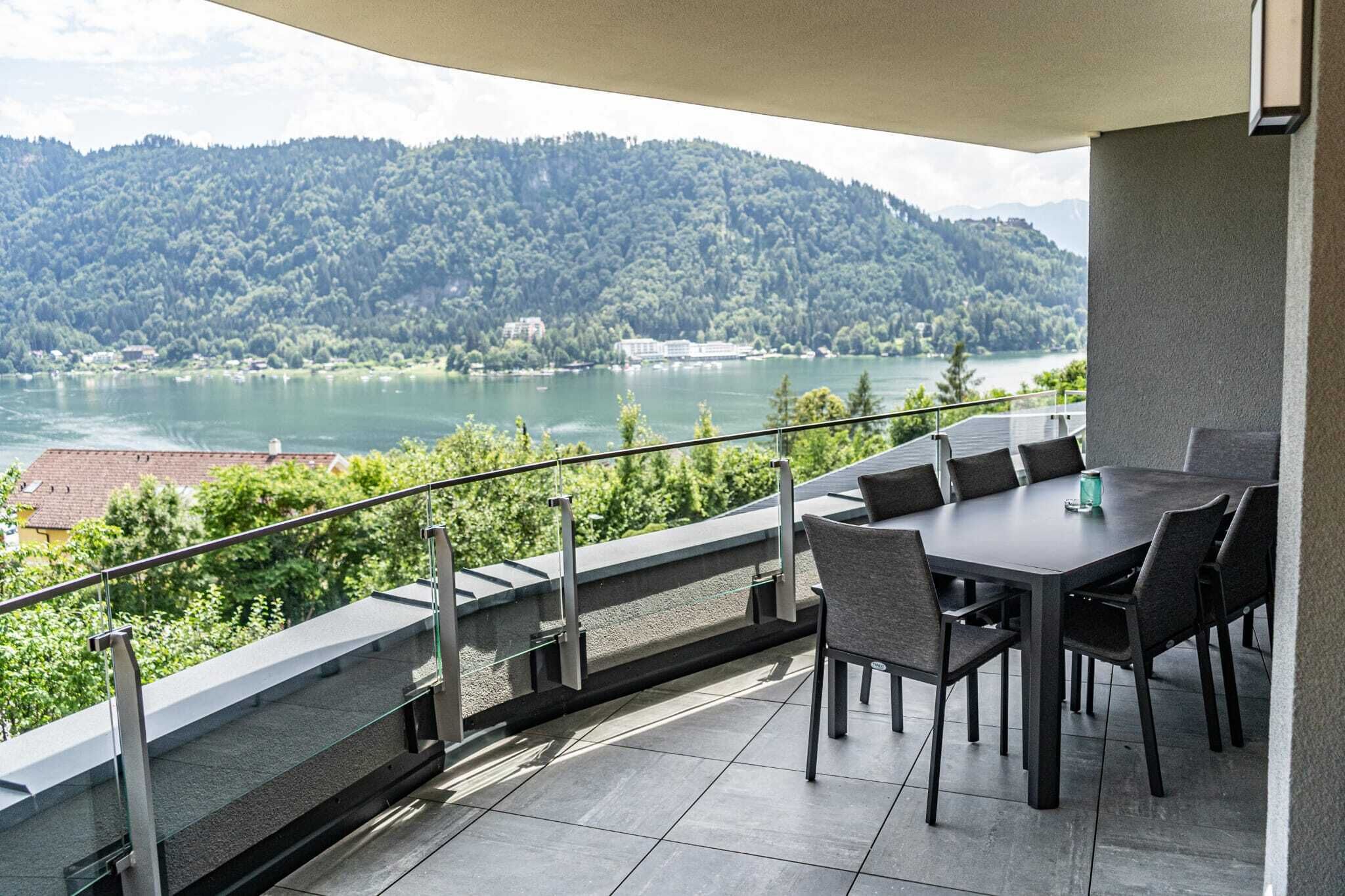 A balcony with a dining table and chairs, overlooking a lake and forested hills.