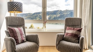 Zwei Sessel mit gemusterten Kissen, eine Stehlampe und Blick auf einen See und Hügel durch ein Fenster.